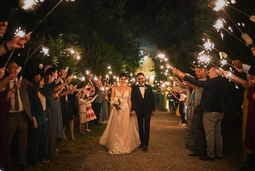 Gli sposi camminano sorridenti lungo un viale notturno mentre amici e familiari, disposti ai lati, tengono in mano stelline luminose creando un corridoio di luce.